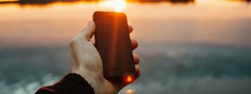 Eine Hand die ein Smartphone hält, im Hintergrund ein Sonnenuntergang über einem See