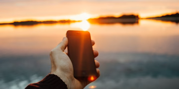 Eine Hand die ein Smartphone hält, im Hintergrund ein Sonnenuntergang über einem See