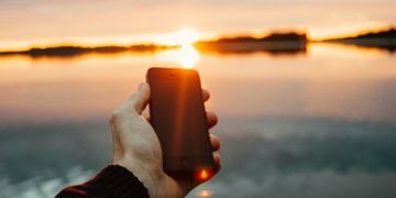 Eine Hand die ein Smartphone hält, im Hintergrund ein Sonnenuntergang über einem See
