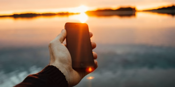 Eine Hand die ein Smartphone hält, im Hintergrund ein Sonnenuntergang über einem See